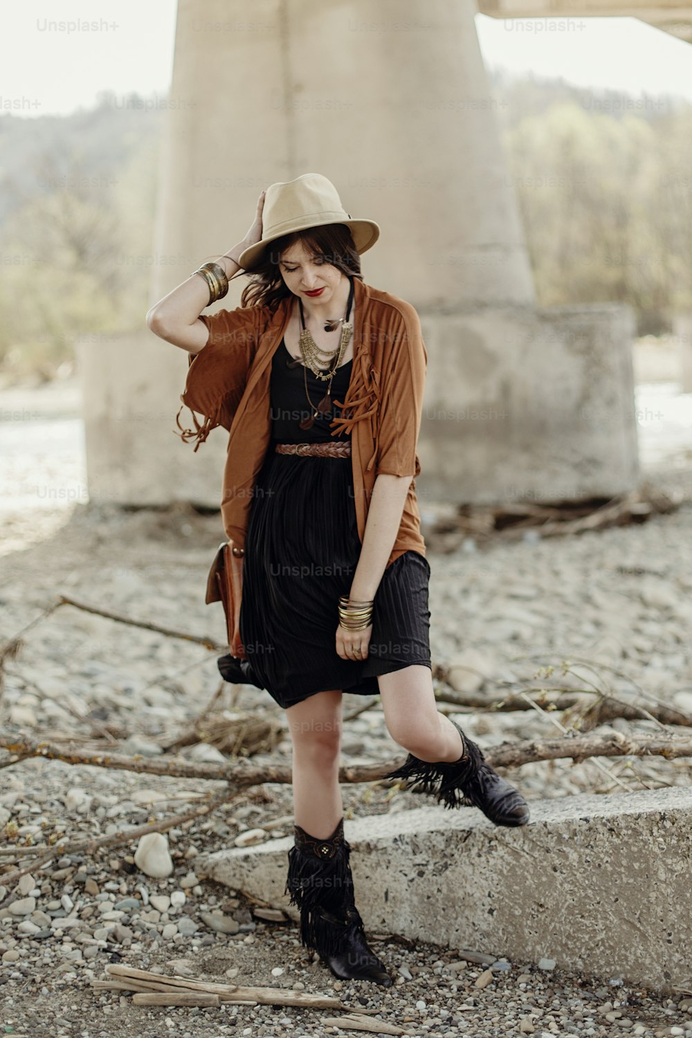 stylish boho woman with jewelry posing at rock wall. beautiful gypsy dressed girl with hat and fringe poncho with sensual look. young girl traveler. fashionable hippie outfit. summer vacation