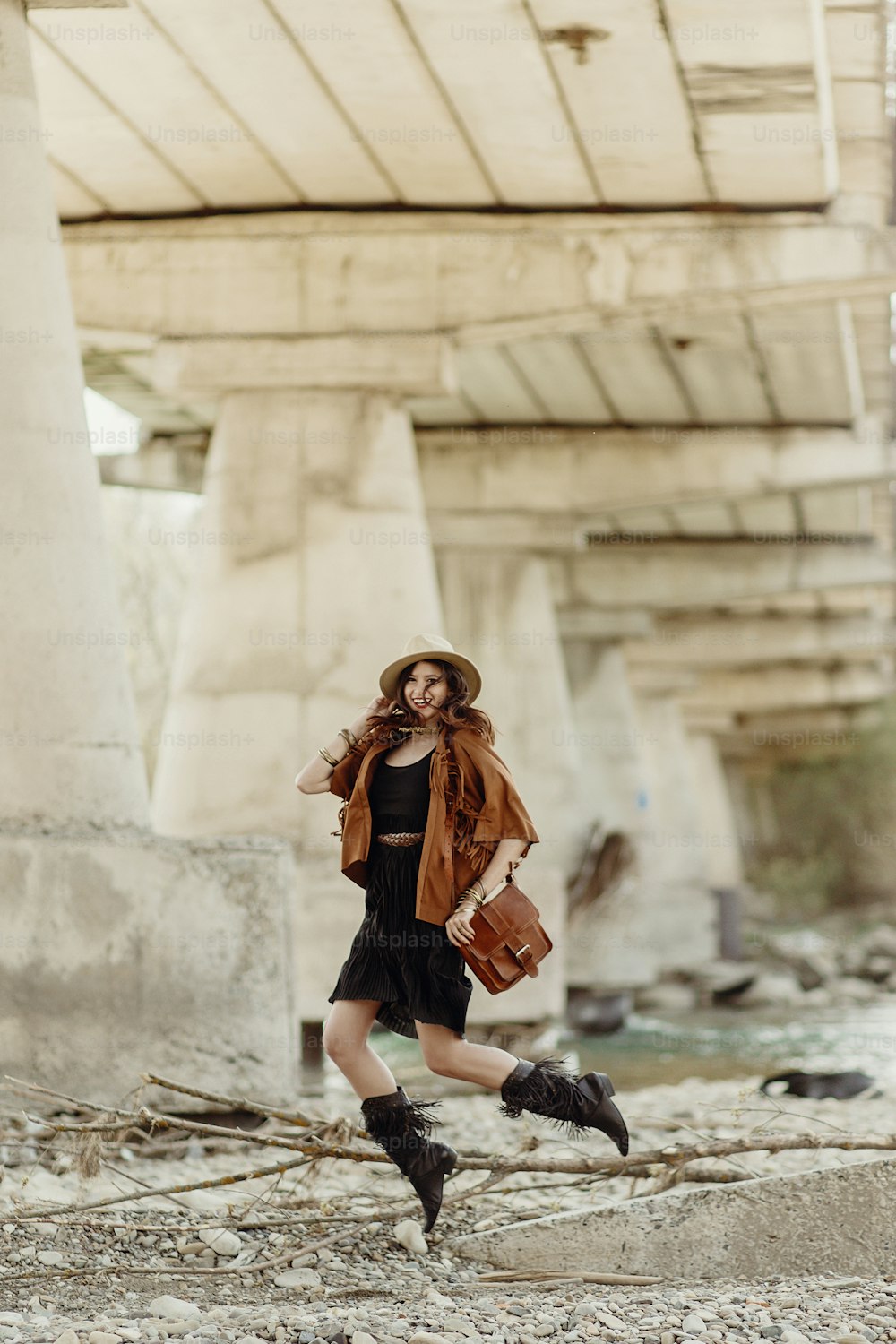 stylish boho woman jumping, having fun, in hat, leather bag, fringe poncho and boots  near river under bridge stone. girl in gypsy hippie look young traveler. joyful moments