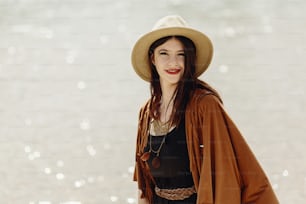 stylish boho traveler woman in hat, fringe poncho posing near water river beach, gypsy hipster girl look, summer travel. atmospheric moment. space for text. wanderlust