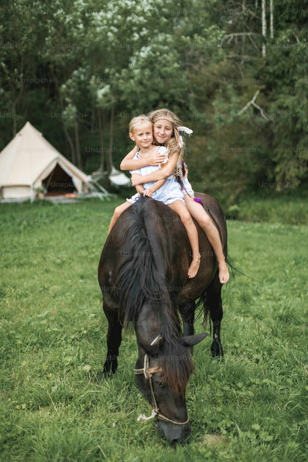 Horseback, two children girls, sisters, riding a horse outdoors, smiling and hugging each other. Girls riding a horse in meadow in summer. Wigwam tents, white teepee tent and trees on the background.