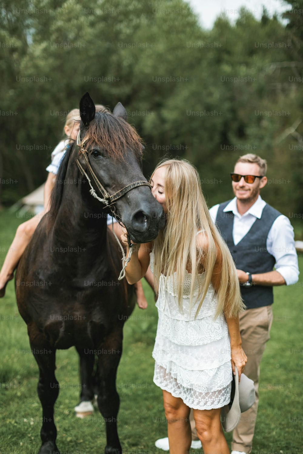 Portrait of a young happy family, parents and children, having fun at countryside outdoors, walking with horse. Kids are riding horse, mother is kissing and petting horse head. Summertime.