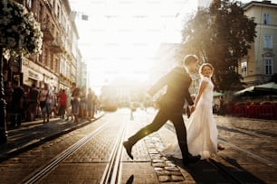 Élégante mariée et le marié courant et souriant dans la rue de la ville européenne le soir. couple de mariage émotionnel de luxe s’amusant. moment sensuel romantique. Des rayons de soleil incroyables