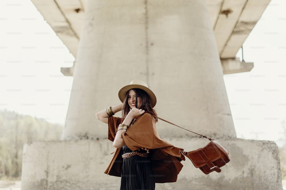 stylish hipster woman having fun, in hat with windy hair near river stones. boho traveler girl in gypsy look dancing with bag. summer travel. atmospheric moment. space for text.