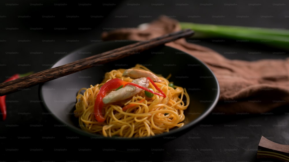 Abgeschnittene Aufnahme von Schezwan Nudeln oder Chow Mein in schwarzer Schüssel mit Essstäbchen auf schwarzem Tisch