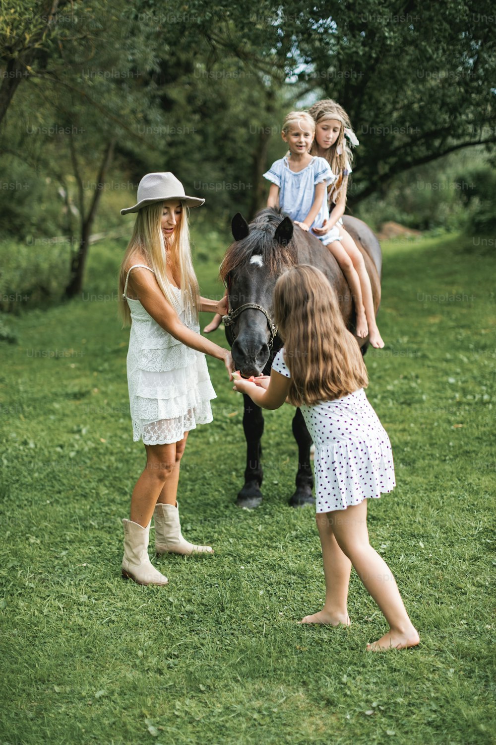 Beautiful young mother in white dress and stylish hat walking with three girls daughters and brown horse outdoors. Two girls riding horse, one sister is feeding the horse with mom. Family with horse.