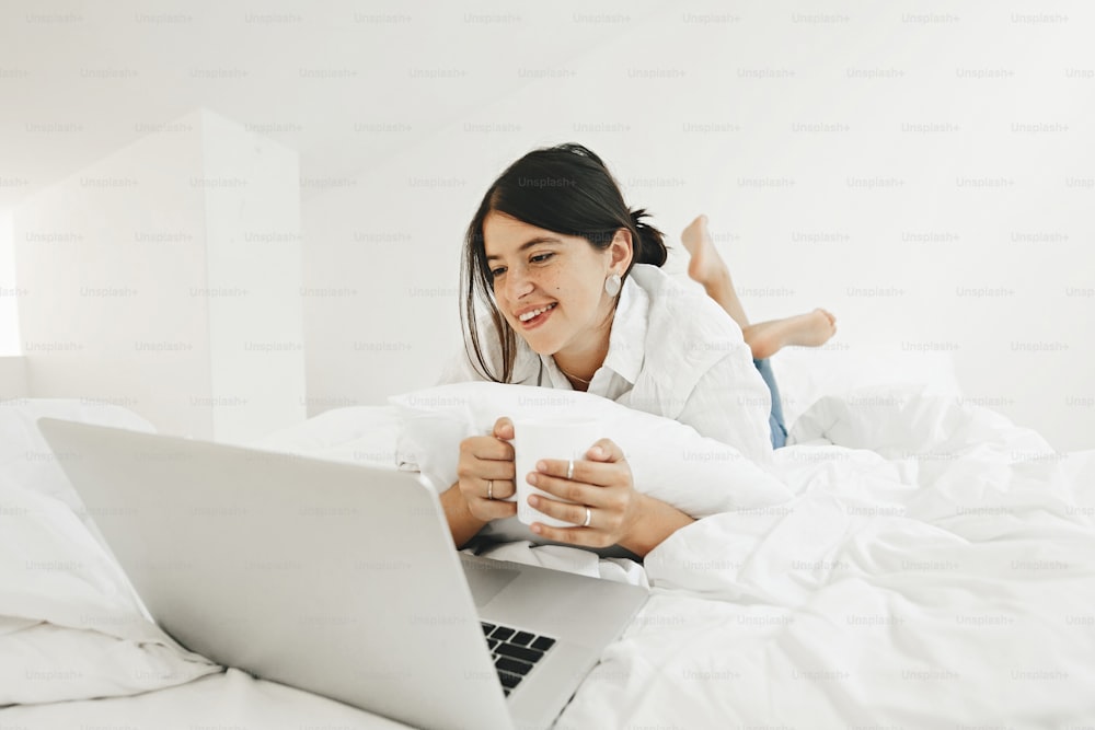 Happy stylish girl looking at laptop, lying on bed in white modern room. Young casual woman shopping online or studying online from home and smiling. Stay home. Freelance