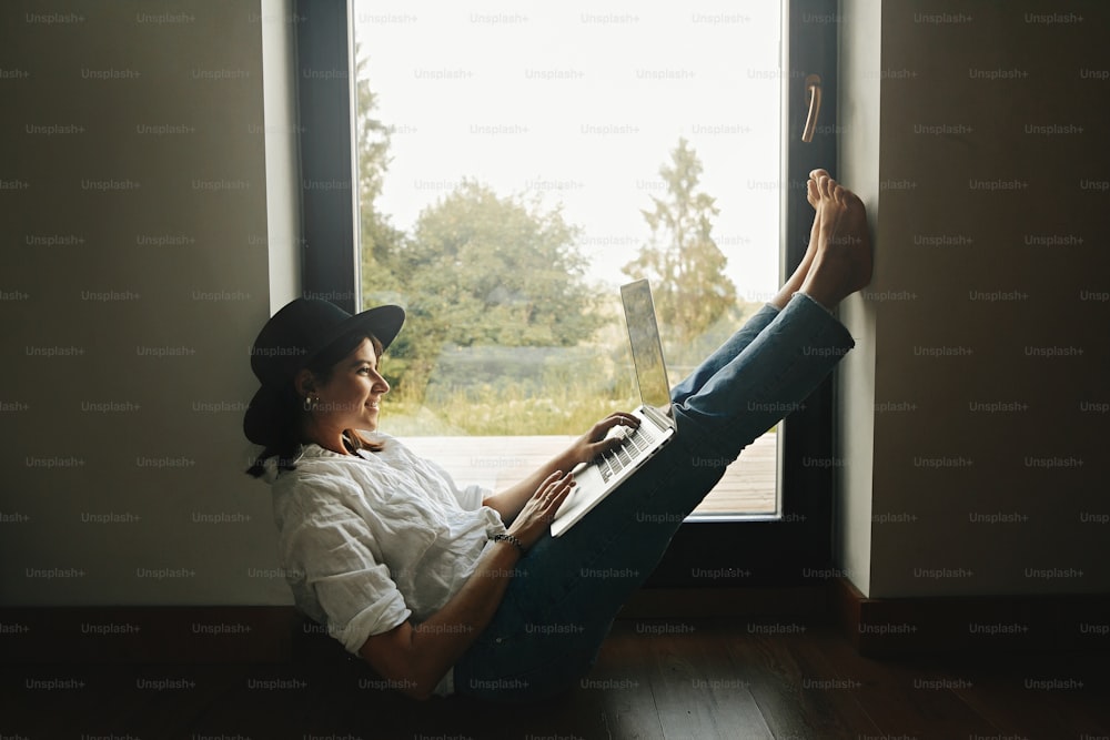 Stylish hipster girl sitting on floor with laptop at big window with view into forest. Young happy woman in hat using laptop, shopping or working online from home. Freelance and freelancer
