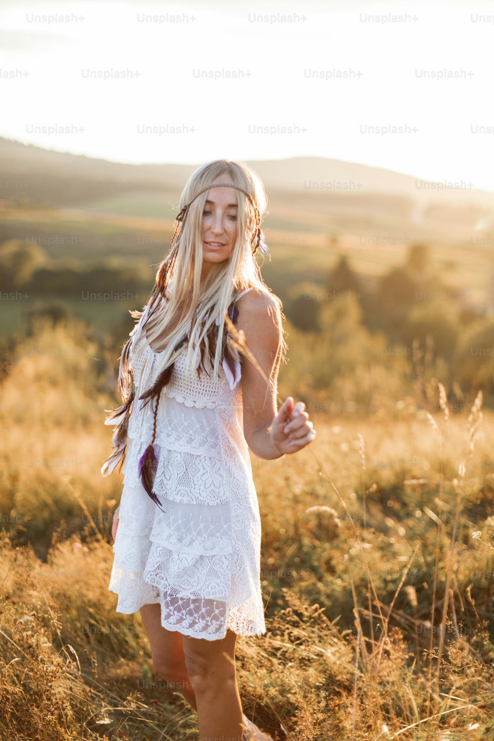 Mujer boho rubia atractiva feliz en vestido blanco y accesorios de plumas en el pelo en el campo de verano al aire libre, puesta del sol. Moda boho, estilo hippie.