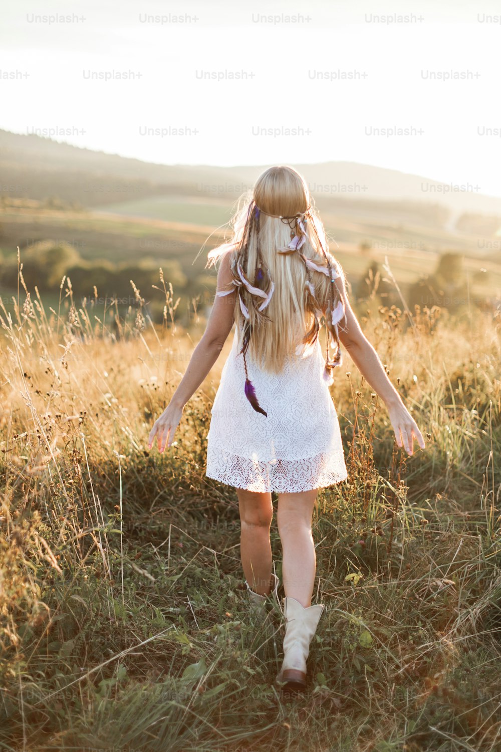 Back view of beautiful young boho woman in white dress with colorful feathers and accessories at sunset in the summer field.