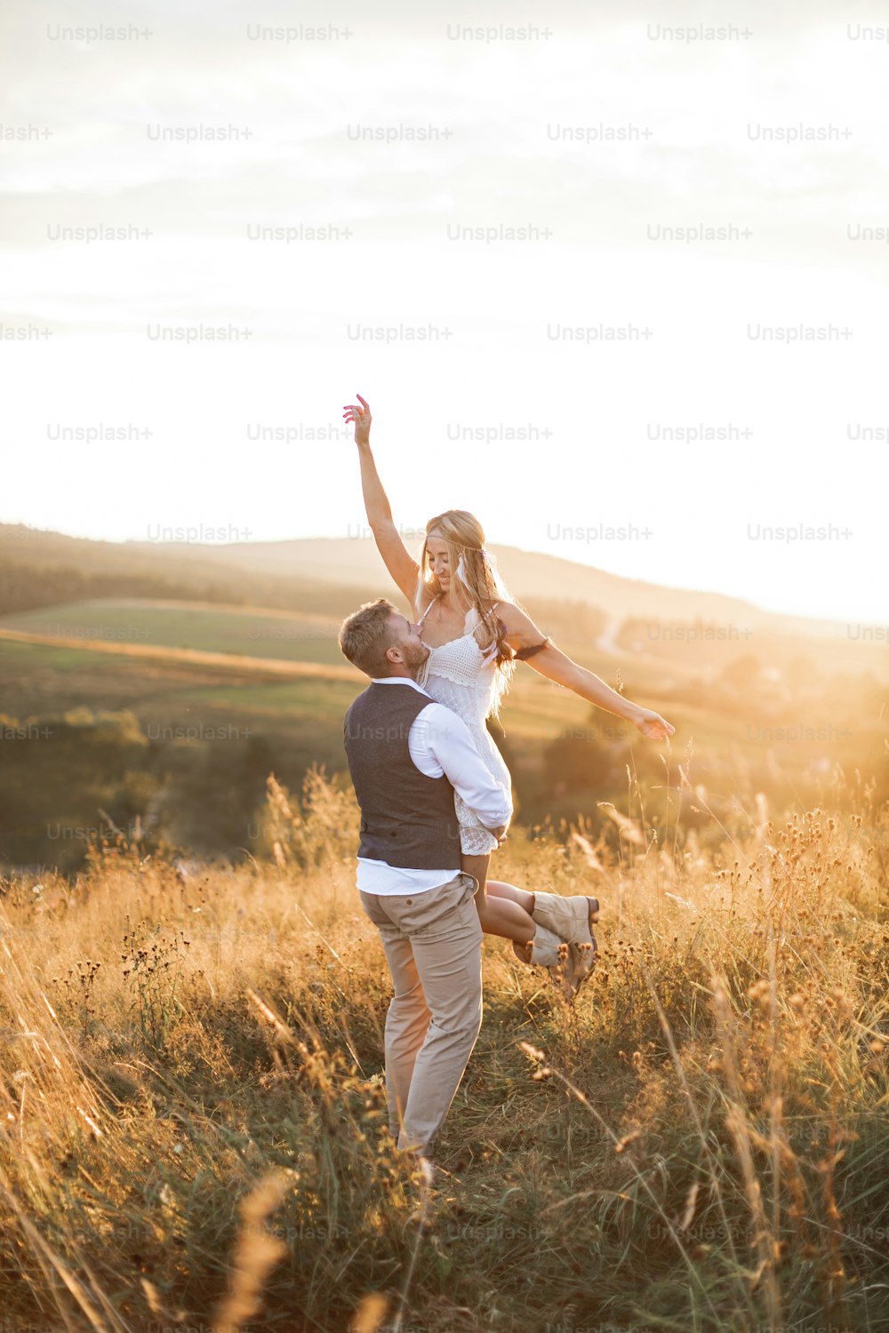 Young couple wearing hipster boho indie style clothes, in love walking in countryside, man holds woman on hands, woman is happy and smiling. Sunny sunset field on background, warm summer day.