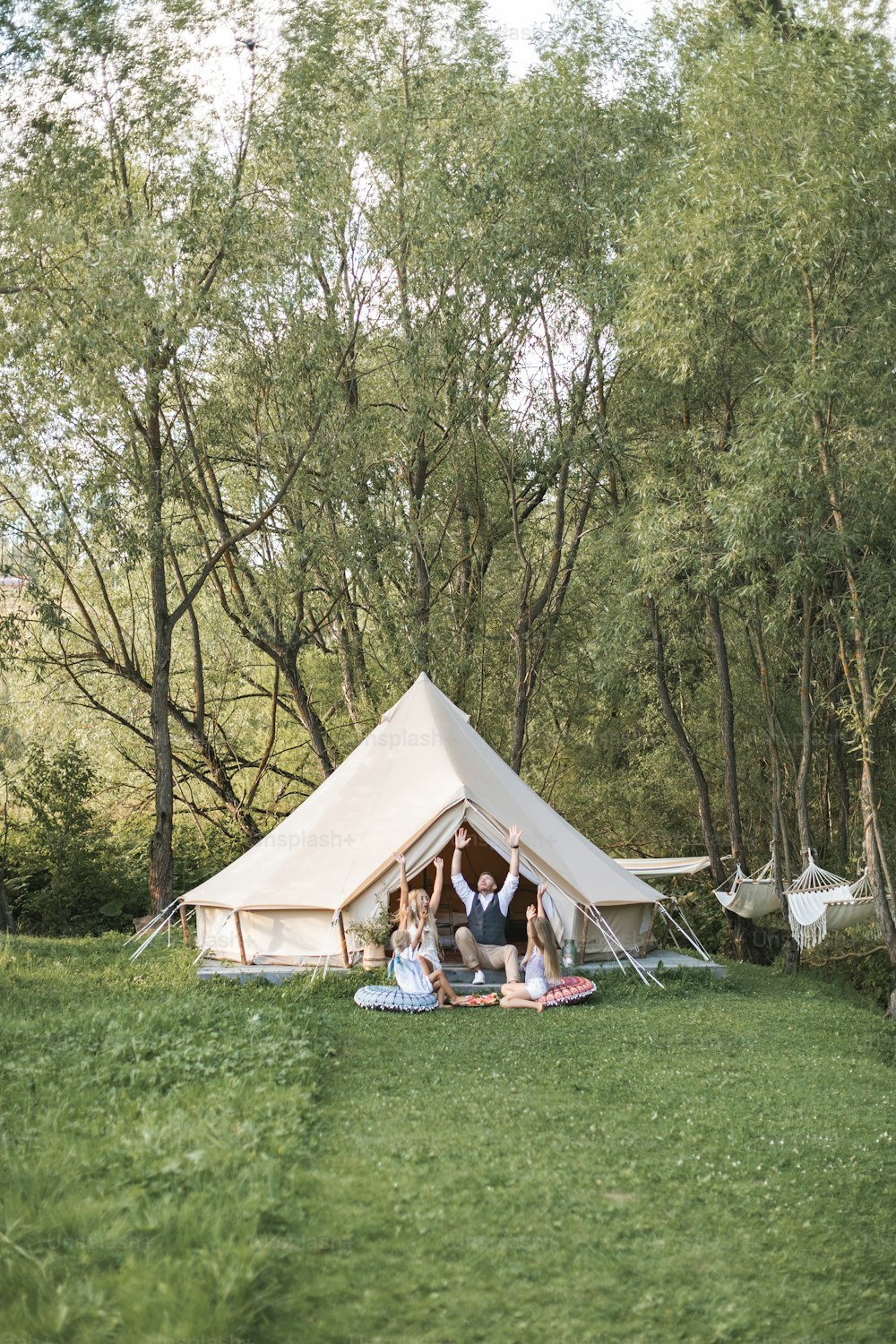 Glückliche junge Familie mit zwei Kindern, die Zeit zusammen in der Natur verbringen, in der Nähe des großen Zeltes sitzen und die Arme hochhalten, lächeln und Spaß haben. Boho rustikales Zelt, Familienkonzept.