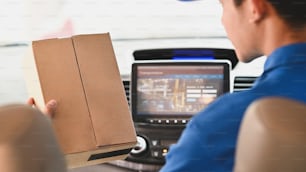 Imagen recortada detrás del repartidor sosteniendo / enviando una caja de cartón al cliente mientras está sentado detrás del volante en la camioneta moderna.