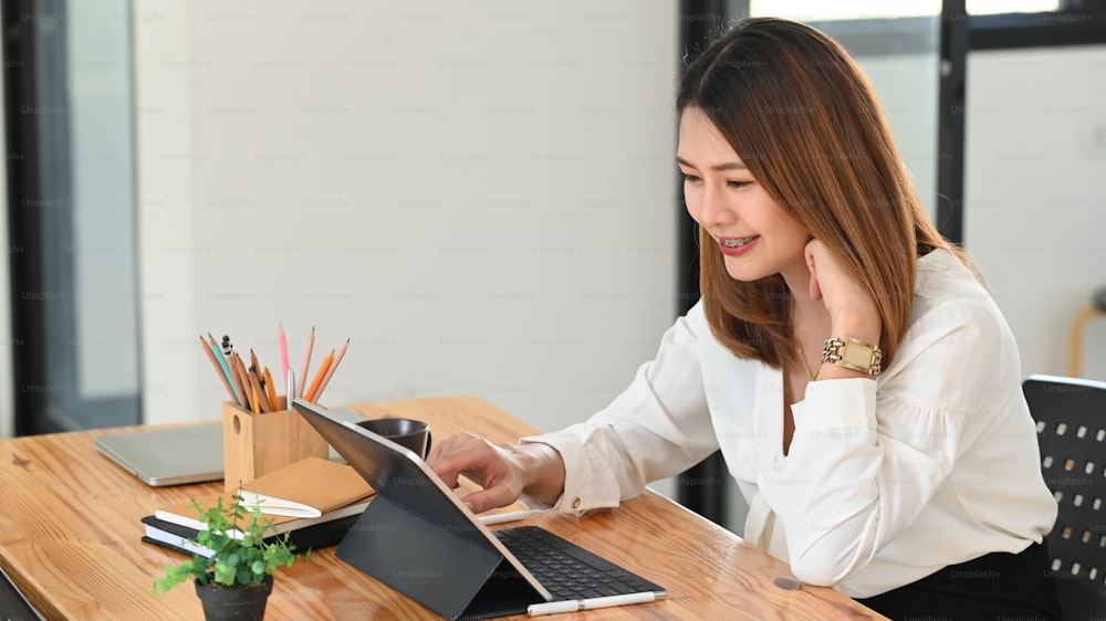 Photo d’une belle jeune femme travaillant comme secrétaire assise à la chaise de bureau et tapant sur l’écran d’une tablette d’ordinateur qui met sur une table en bois avec une plante en pot, un porte-crayon, un cahier.