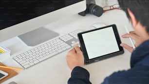 Abgeschnittene Aufnahme eines intelligenten Mannes, der den Stift in den Händen hält, während er ein weißes Computertablett mit leerem Bildschirm verwendet und am modernen Arbeitstisch mit komfortablem Arbeitsplatz als Hintergrund sitzt.