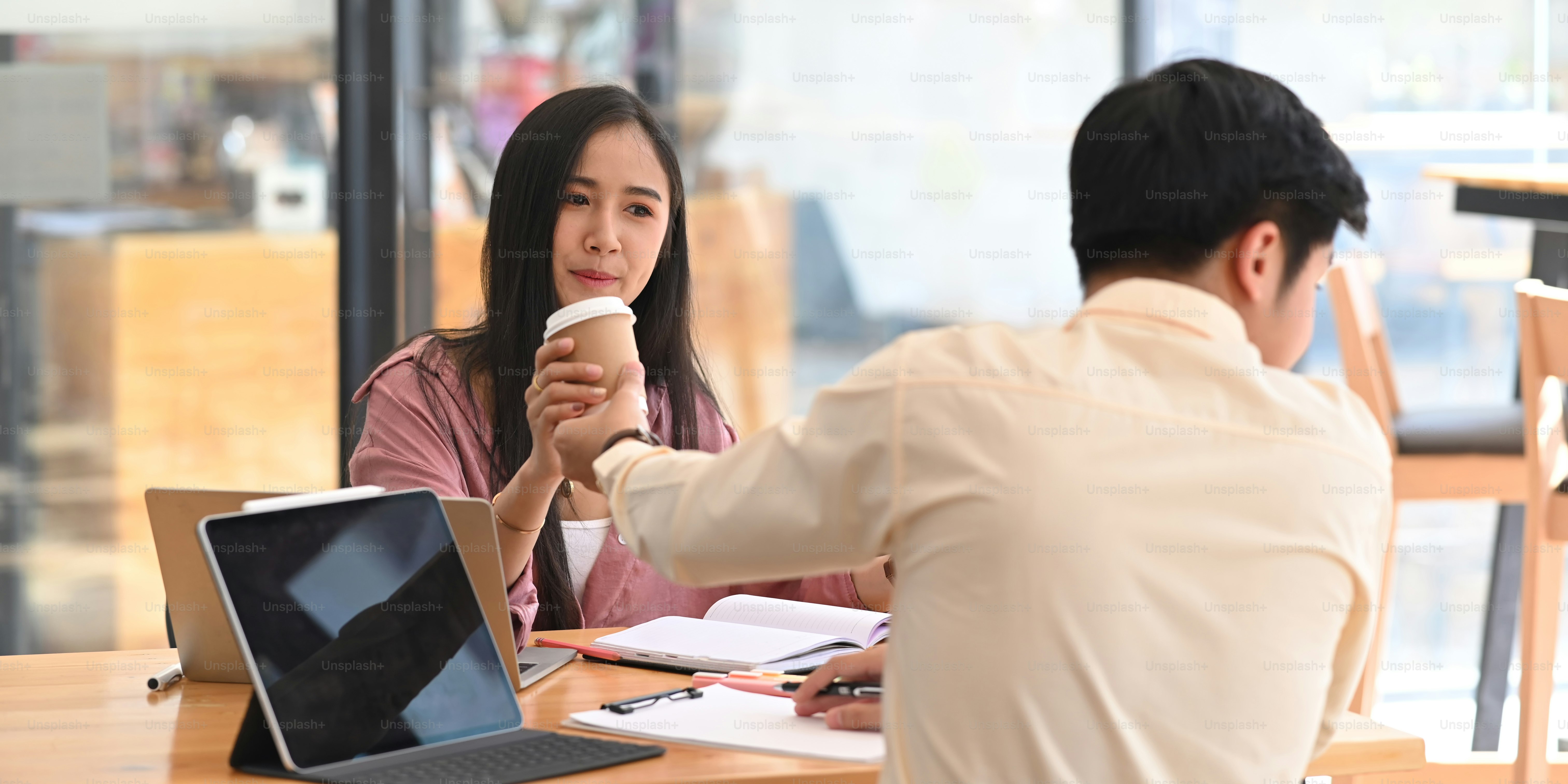 Photo of smart businessman sending a coffee cup to his colleague while sitting at the wooden meeting table with computer laptop, business document and tablet with orderly office as background.