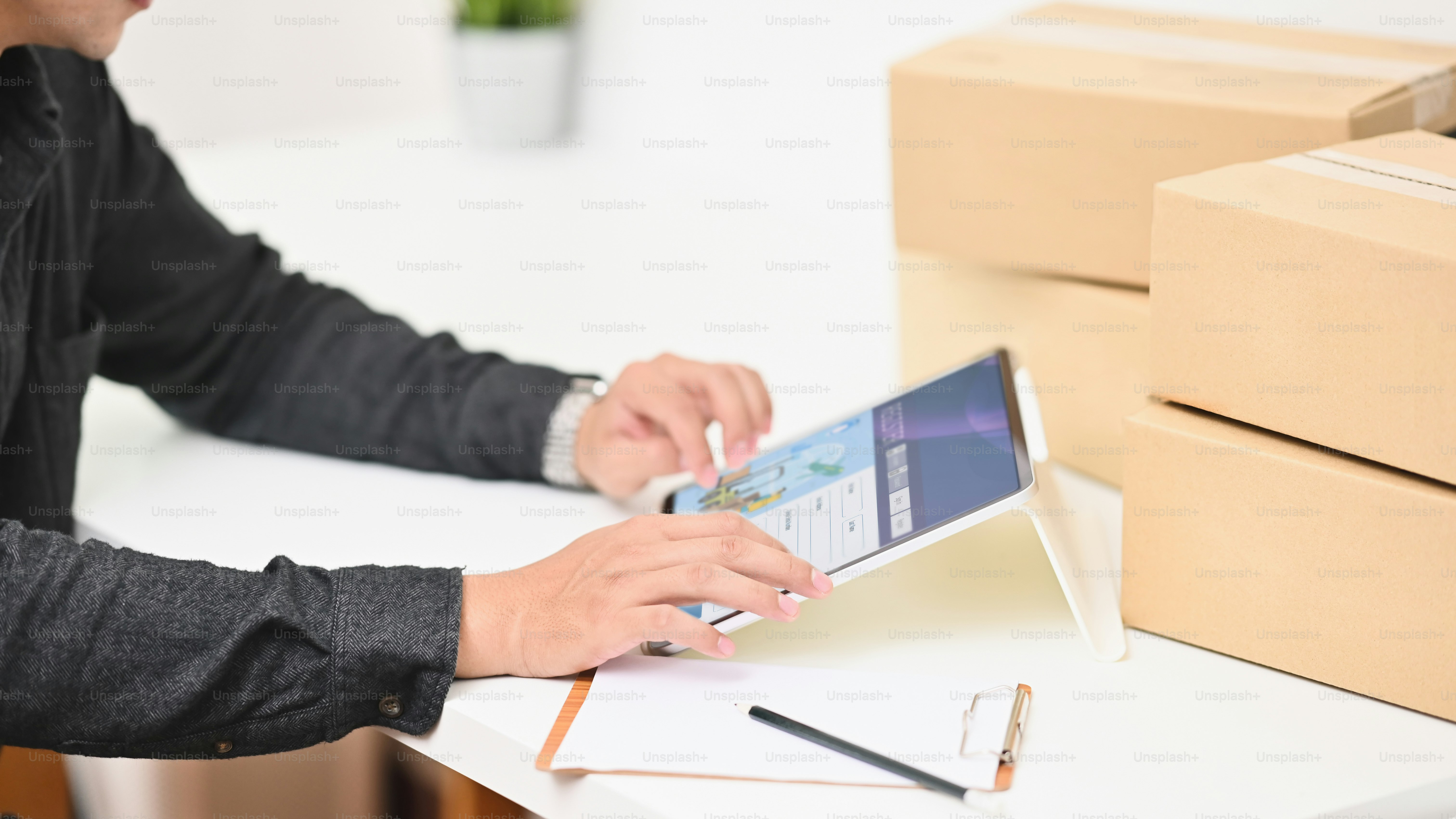 Portrait of young business owner using a computer tablet while sitting at the modern working desk surrounded by stack of cardboard boxes with comfortable living room as background.