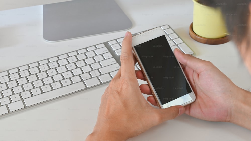 Cropped shot of smart man holding white smartphone with black blank screen in hands while sitting at the modern working table with comfortable workplace as background.
