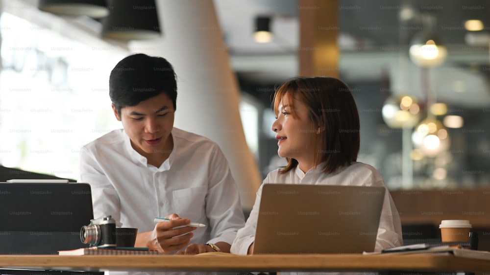Photo of young woman and man in white shirt working as financial developer team working together while sitting at the modern wooden table with luxury office as background.