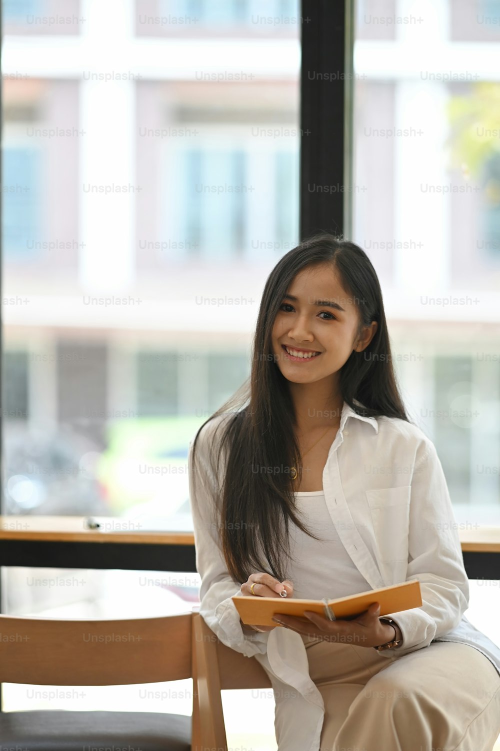 Lächelnde schöne Frau im weißen Hemd, die ein Notizbuch in den Händen hält, während sie an der modernen Theke im Café mit Glaswand als Hintergrund sitzt.