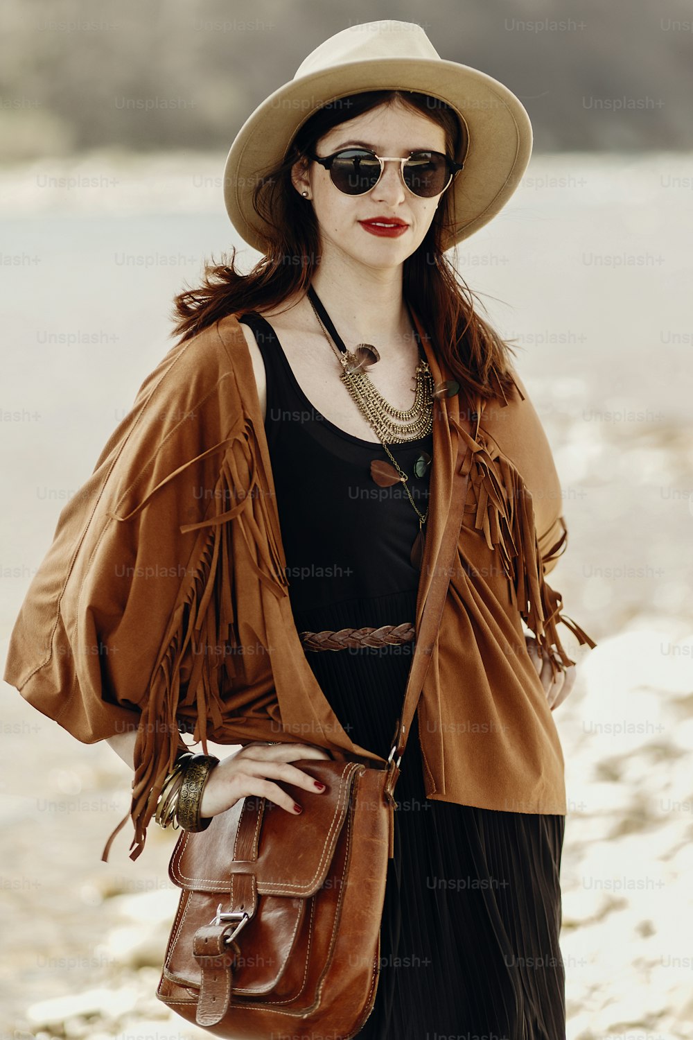 stylish hipster boho woman in sunglasses with hat, leather bag, fringe poncho and accessory. traveler girl look, near beach in mountains. atmospheric moment. summer travel.