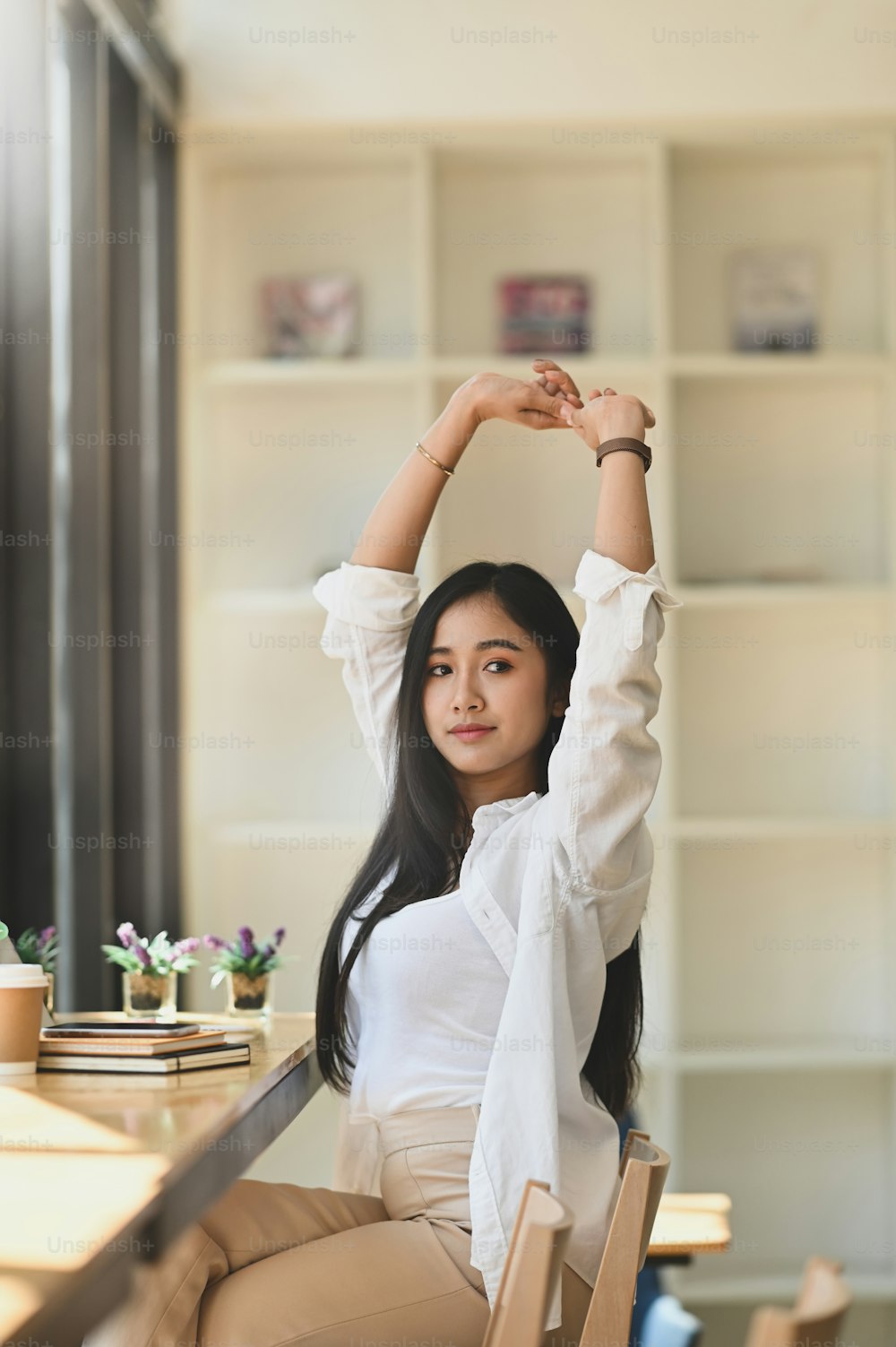 Photo of beautiful woman in white shirt that working as secretary stretching her hands while sitting at the modern wooden counter bar with cafe book shelf as background. Relaxing girl concept.