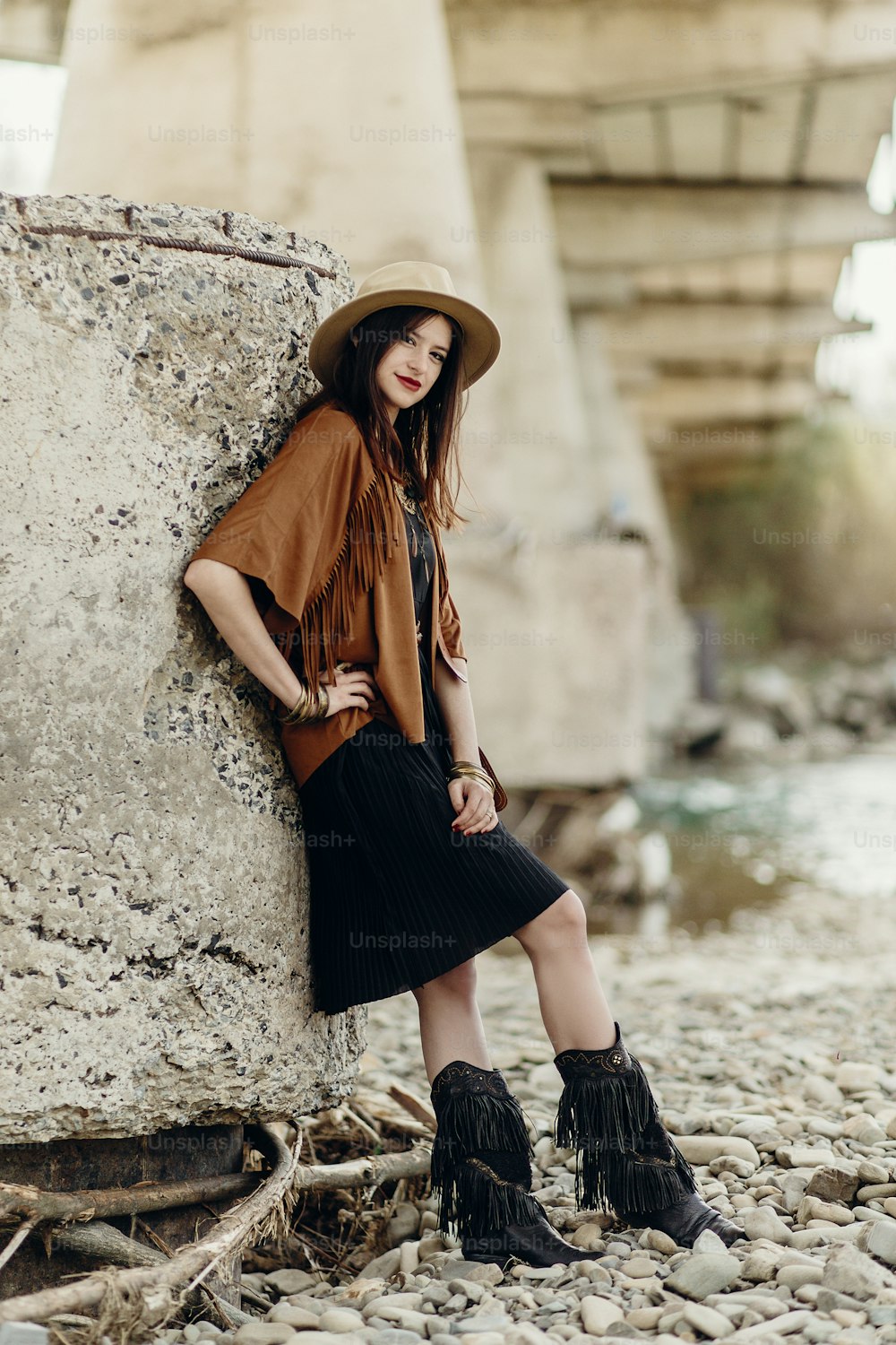 beautiful stylish boho woman with hat, fringe poncho and boots. girl in gypsy hippie look young traveler posing near river rocks in mountains. sensual look. atmospheric moment. space for text