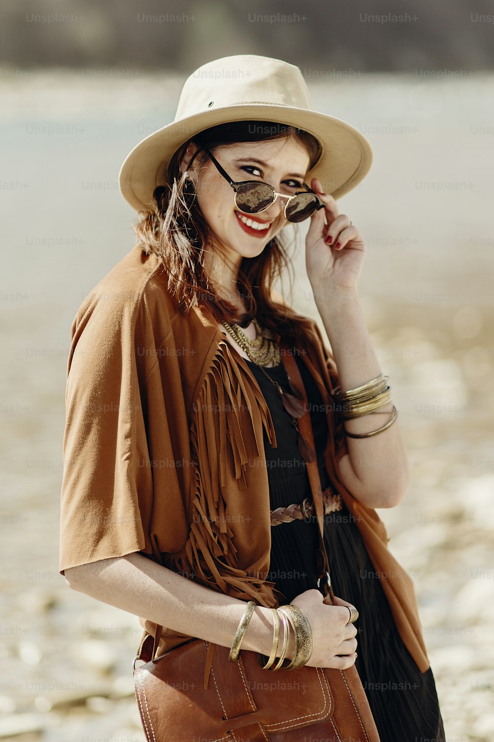 stylish hipster boho woman smiling in sunglasses with hat, leather bag, fringe poncho and accessory. traveler girl look, near beach in mountains. atmospheric moment. summer travel.