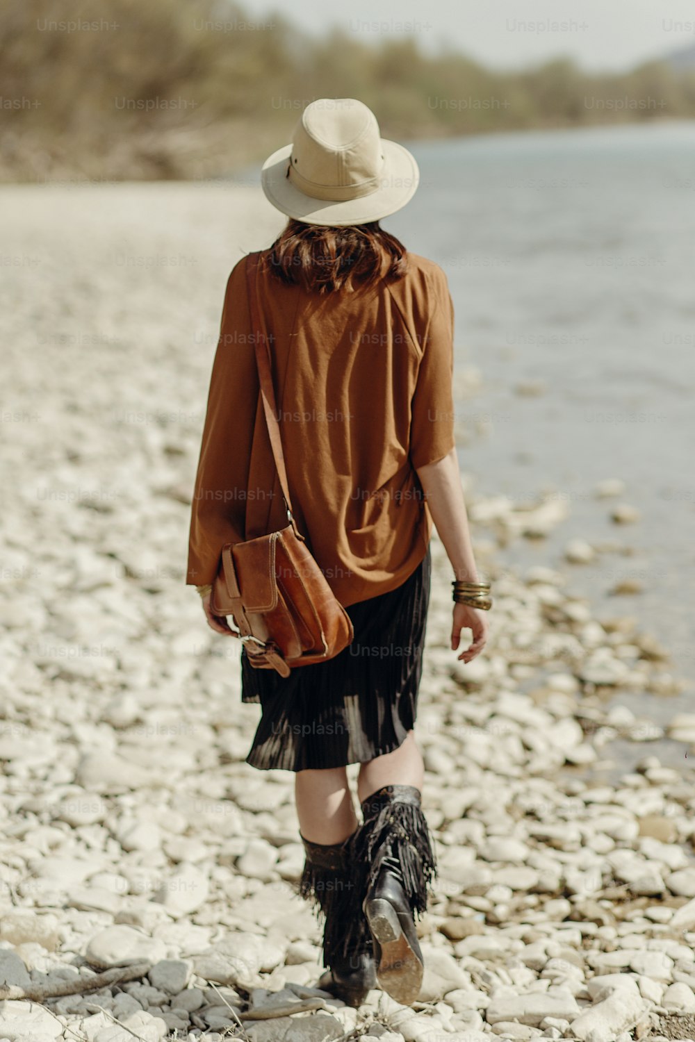 stylish boho gypsy woman in hat, fringe poncho and boots walking on river beach,back view. hipster traveler girl look, summer travel. atmospheric moment. space for text. wanderlust
