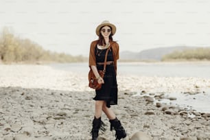 stylish boho traveler woman in hat, fringe poncho walking near water river beach in mountains, gypsy hipster girl. wanderlust summer travel. atmospheric moment. space for text.