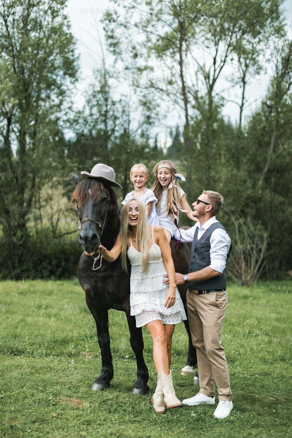 Happy family, parents and children, enjoying the horse presence. Girls are riding the horse, wearing hat. Man and woman parents having fun and laughing. Countryside, horse riding outdoors.