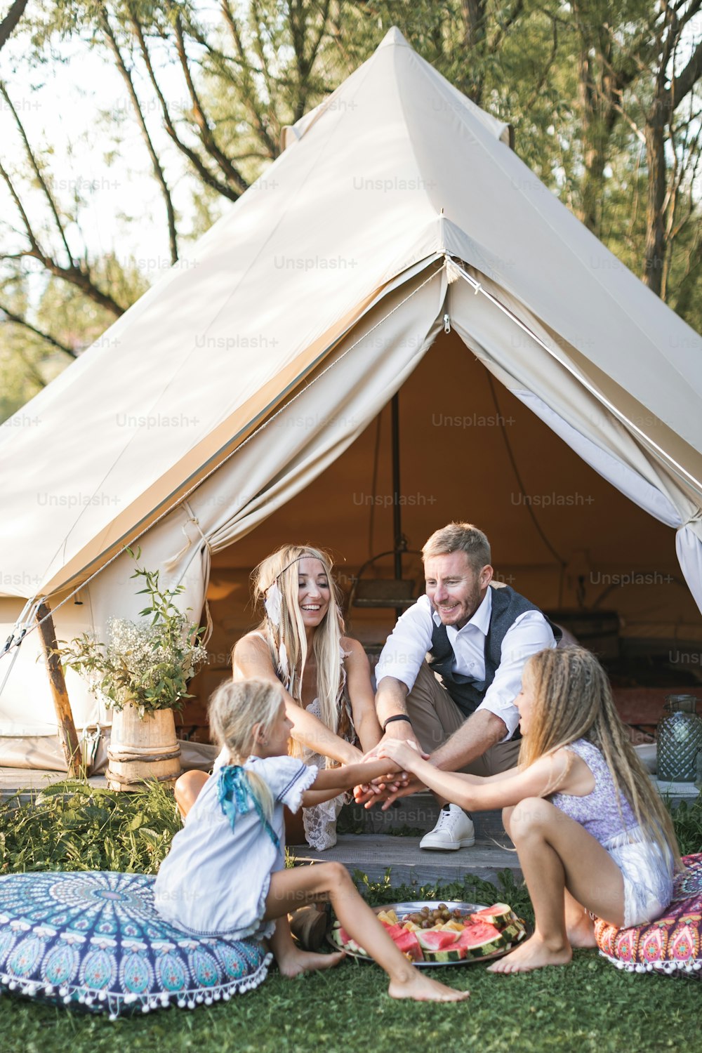 Happy cheerful family, father, mother and two daughters, putting their hands together, while sitting near the big wigwam tent with boho decorations and pillows on a sunny day outdoors.