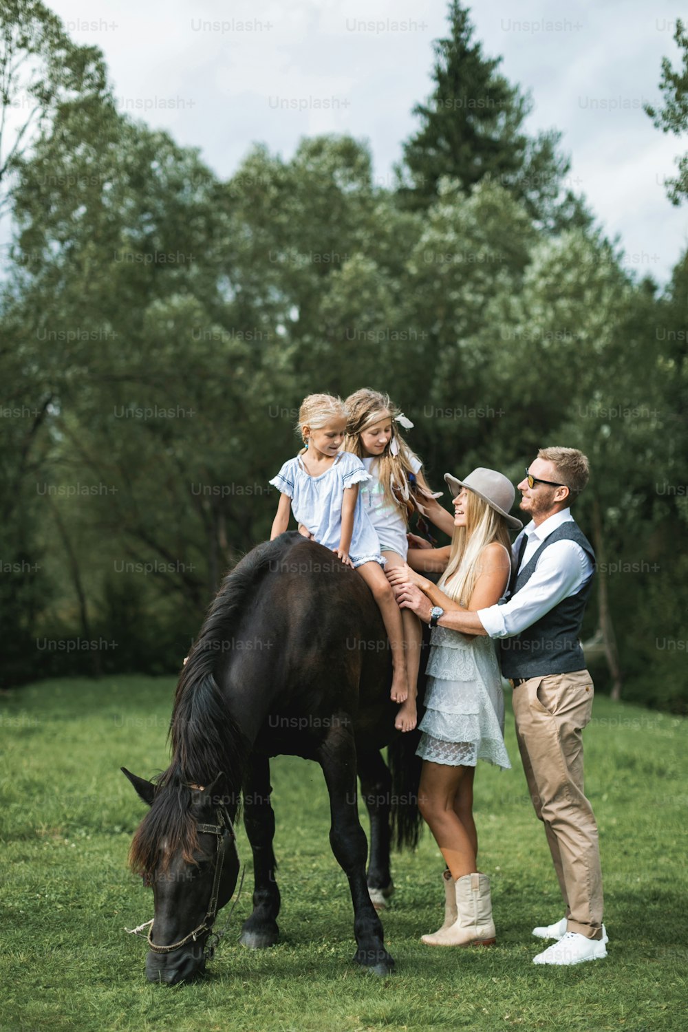 Young stylish family in cowboy wear, father, mother teaching their two little daughters to ride a horse. Family horse riding on the nature in countryside.