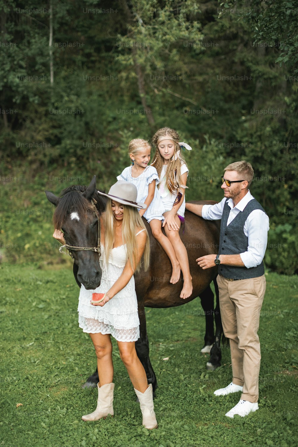 Happy family, father, mother and two children girls riding horse, relaxing in green meadow. Family, wearing stylish boho, ranch clothes, walking with horse outdoors.