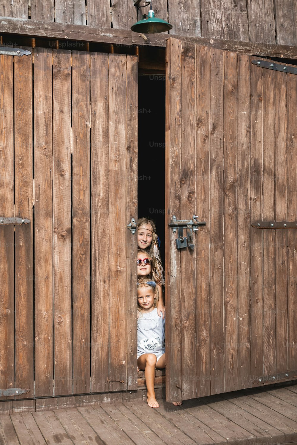 Pequeñas tres chicas de campo con ropa boho informal, sonriendo mientras están sentadas en la puerta de un granero de madera en el campo o la granja, caluroso día de verano. Niños divirtiéndose en granero de madera.