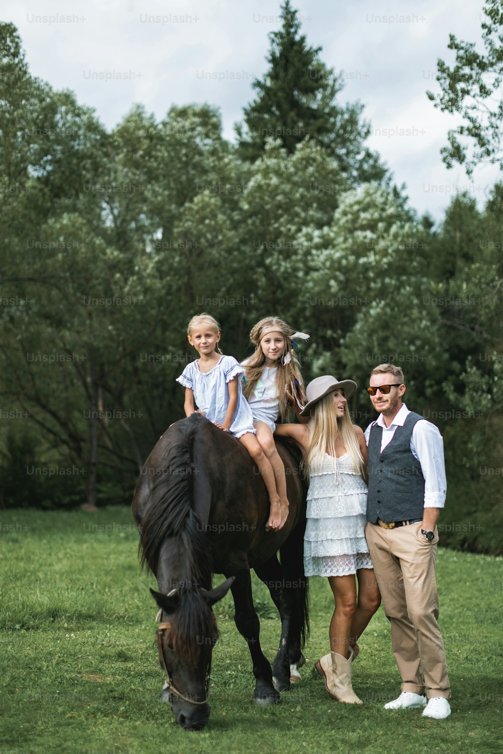 Family walk, horse riding concept. Two little girls riding a brown horse, while young stylish parents in casual cowboy wear standing near horse and looking each other. Hppy family riding horse.