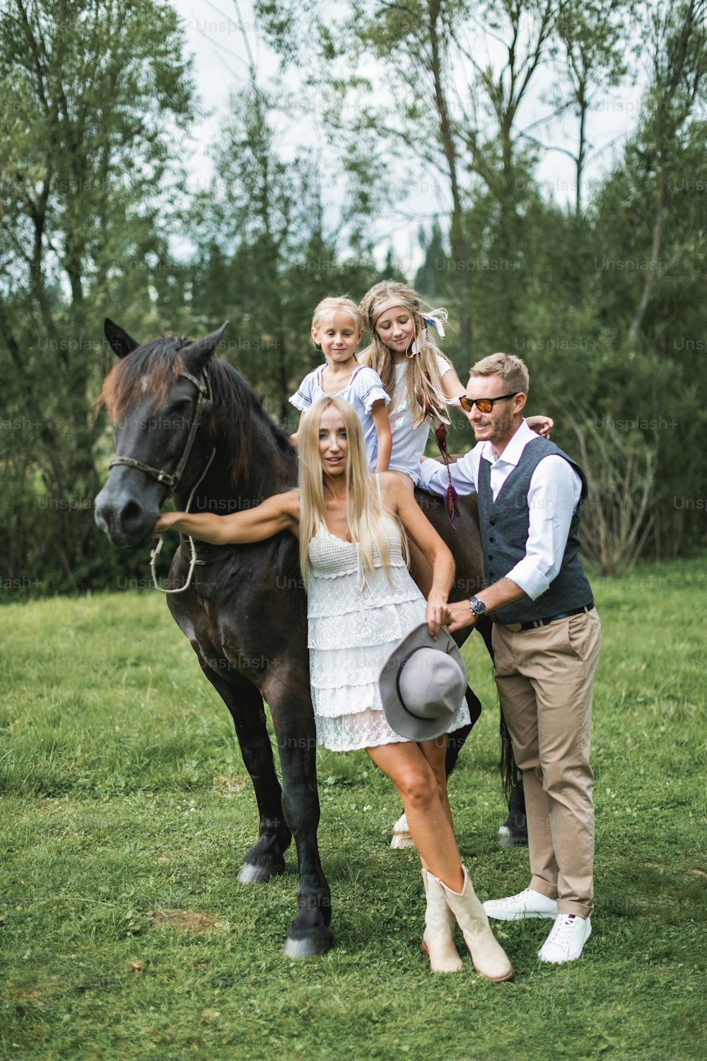 Cute two girls sisters riding horse on nature background, having walk with their mother and father in stylish boho cowboy wear, looking at camera and smiling. Stylish young family walking with horse.