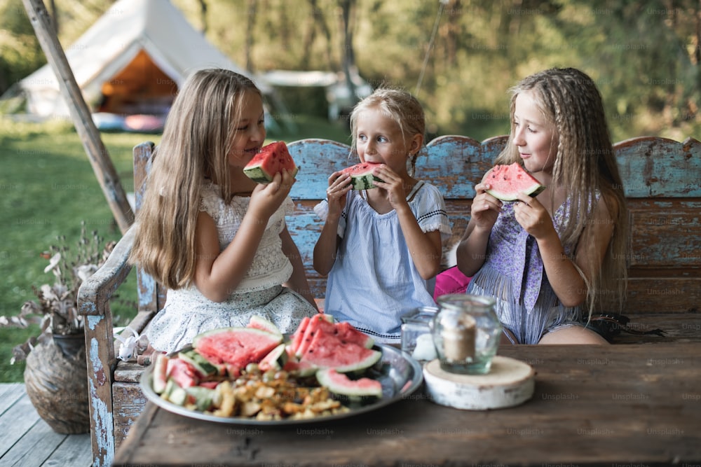 Three cute happy smiling girls, sisters, fiends, sitting at the table on vintage wooden bench and eating watermelon outdoors, in tent, stylish boho wigwam on background. Countryside summer holidays.