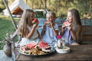 세 명의 귀여운 행복한 미소 짓는 소녀, 자매, 악마, 빈티지 나무 벤치의 테이블에 앉아 야외에서 수박을 먹고 텐트에서 세련된 보헤미안 가발을 배경으로 하고 있습니다. 시골 여름 휴가.