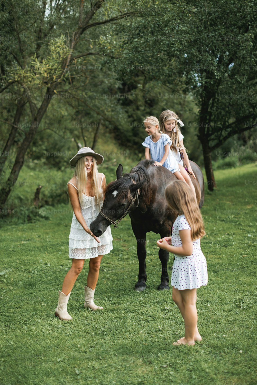 Beautiful young mother in white dress and stylish hat walking with three girls daughters and brown horse outdoors. Two girls riding horse, one sister is feeding the horse with mom. Family with horse.