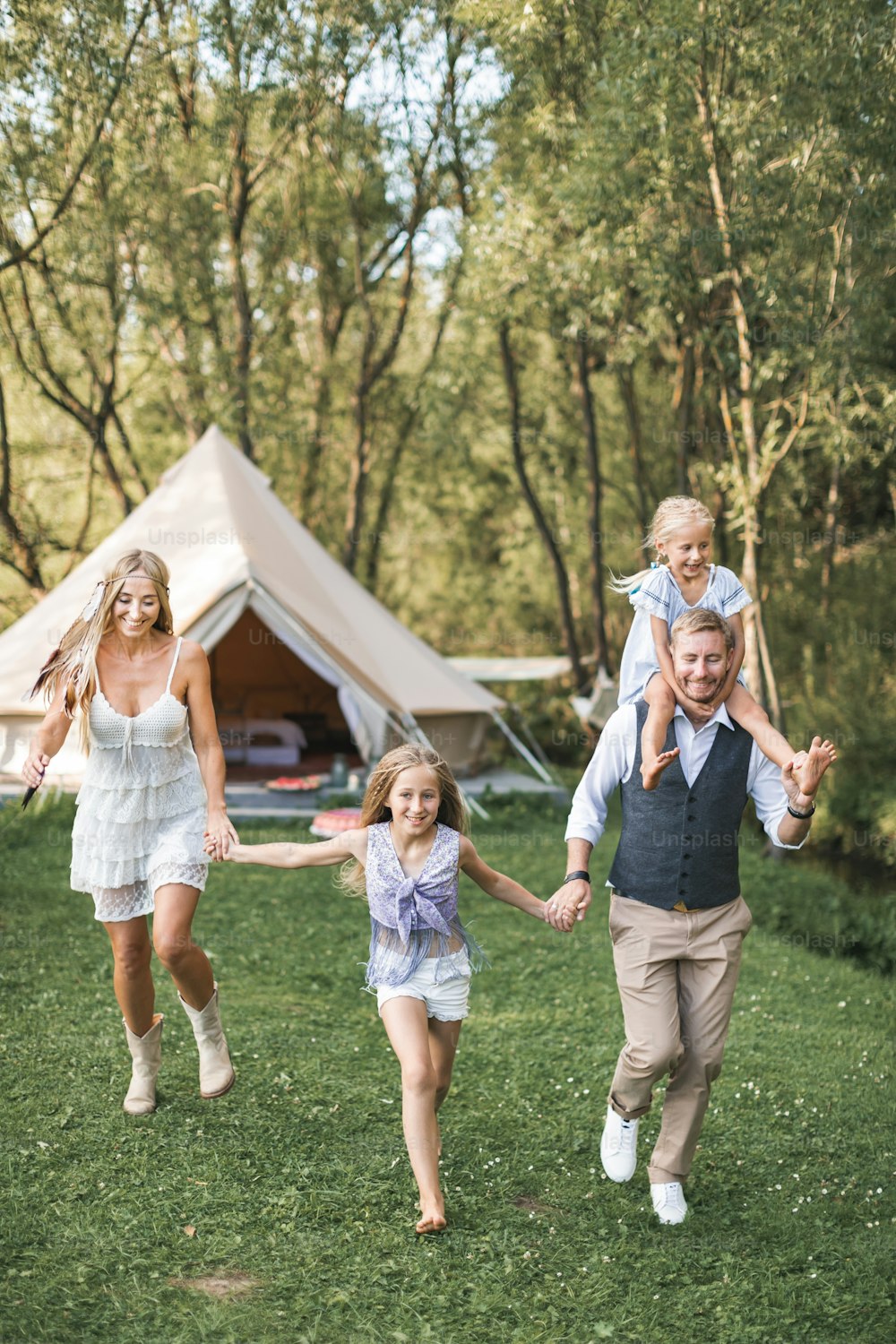 Young family in boho casual wear, dad, mom and two daughters holding hands and running and jumping in a green park, big wigwam tent on the background. Family walk, outdoors.
