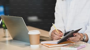 Imagen recortada de cintura para arriba de la mujer de negocios tomando notas/escribiendo en el cuaderno que pone en la mesa de trabajo blanca con la taza de café, la computadora portátil y la planta en maceta sobre la oficina como fondo.