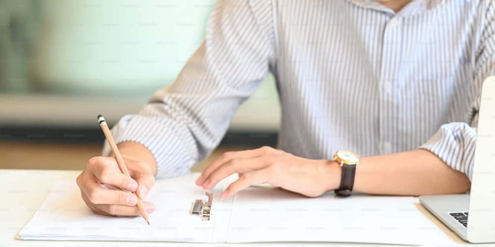 줄무늬 셔츠를 입은 사업가가 사무실을 배경으로 현대적인 작업 테이블에 앉아 문서 파일에 글을 쓰고 있는 모습.