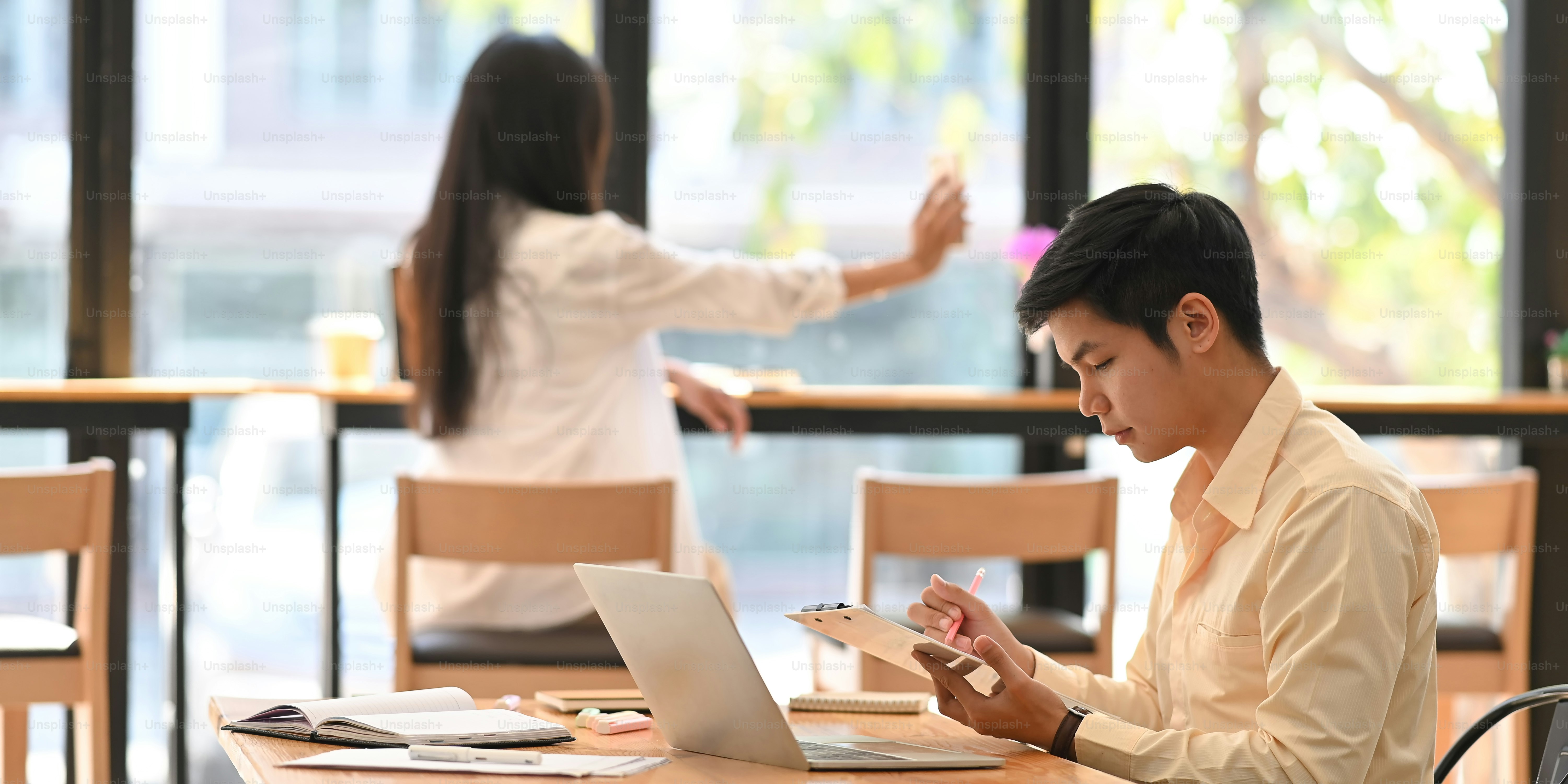 Side shot image of smart businessman in white shirt writing on clipboard while sitting at the wooden working table with cafe/restaurant as background.