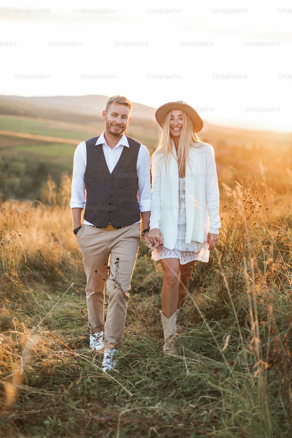Homem bonito e elegante de camisa, colete e calça e mulher bonita boho no vestido, jaqueta e chapéu andando no campo com fardos de palha, de mãos dadas e sorrindo. Noite de verão, pôr do sol.