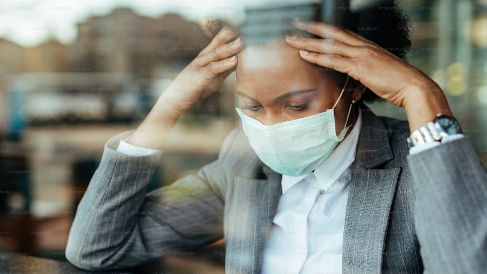 Donna d'affari afroamericana preoccupata che indossa una maschera facciale e pensa mentre si tiene la testa per il dolore. La vista è attraverso il vetro.