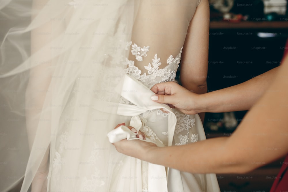 ウェディングドレスを着て美しいブルネットの花嫁、花嫁介添人が花嫁がホテルの部屋で結婚式のコルセットをクローズアップするのを手伝う、準備のコンセプト