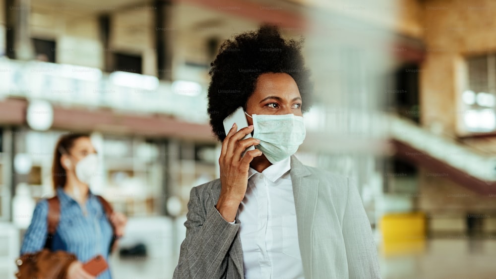 Empresaria afroamericana con máscara protectora mientras se comunica por teléfono móvil en el aeropuerto.