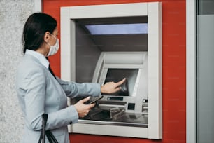 防護マスクを着用したエレガントなビジネスウーマンが街の通りに立ち、ATM機を使って現金を引き出します。コロナウイルスまたはCovid-19ウイルスのパンデミックのコンセプト。