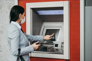 Elegant business woman with protective mask standing on city street and using ATM machine to withdraw cash. Corona or Covid-19 virus pandemic concept.