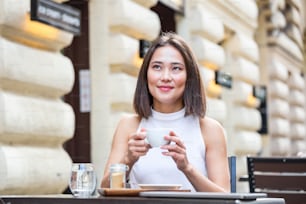 웃는 아시아 여자는 커피를 마시고 휴대폰을 사용하고 있다. 커피 한 잔을 즐기는 만족스러운 여성. 커피숍에서 하얀 머그잔에서 커피를 마시는 아름다운 소녀의 초상화를 닫습니다.